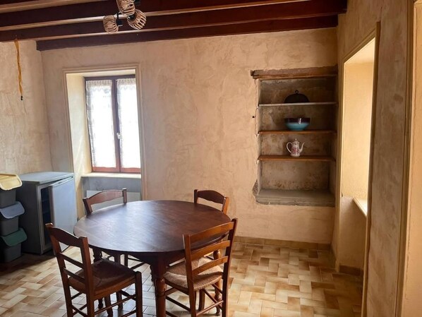 A dining area with a round wooden table and chairs, a window with curtains, and a shelf with kitchenware.