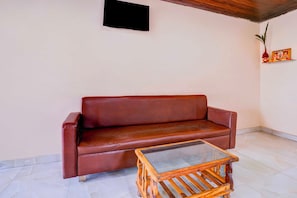 A room with a brown leather sofa, a glass-top wooden coffee table, a wall-mounted flat-screen TV, and a small shelf with a framed picture. - Collection O Khoni Taloja Bypass (Mumbai)