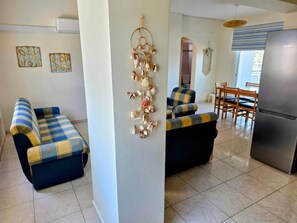 A living room with a sofa, a wall decor, a dining area with chairs, and a refrigerator. - Niki’s Coastal Escape | 2-Bedroom Stay near Mackenzie Beach (Larnaka)