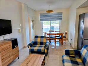 Ein Wohnzimmer mit einer Sofagarnitur, einem Holztisch, einem Fernseher und einem Esstisch mit Stühlen.
