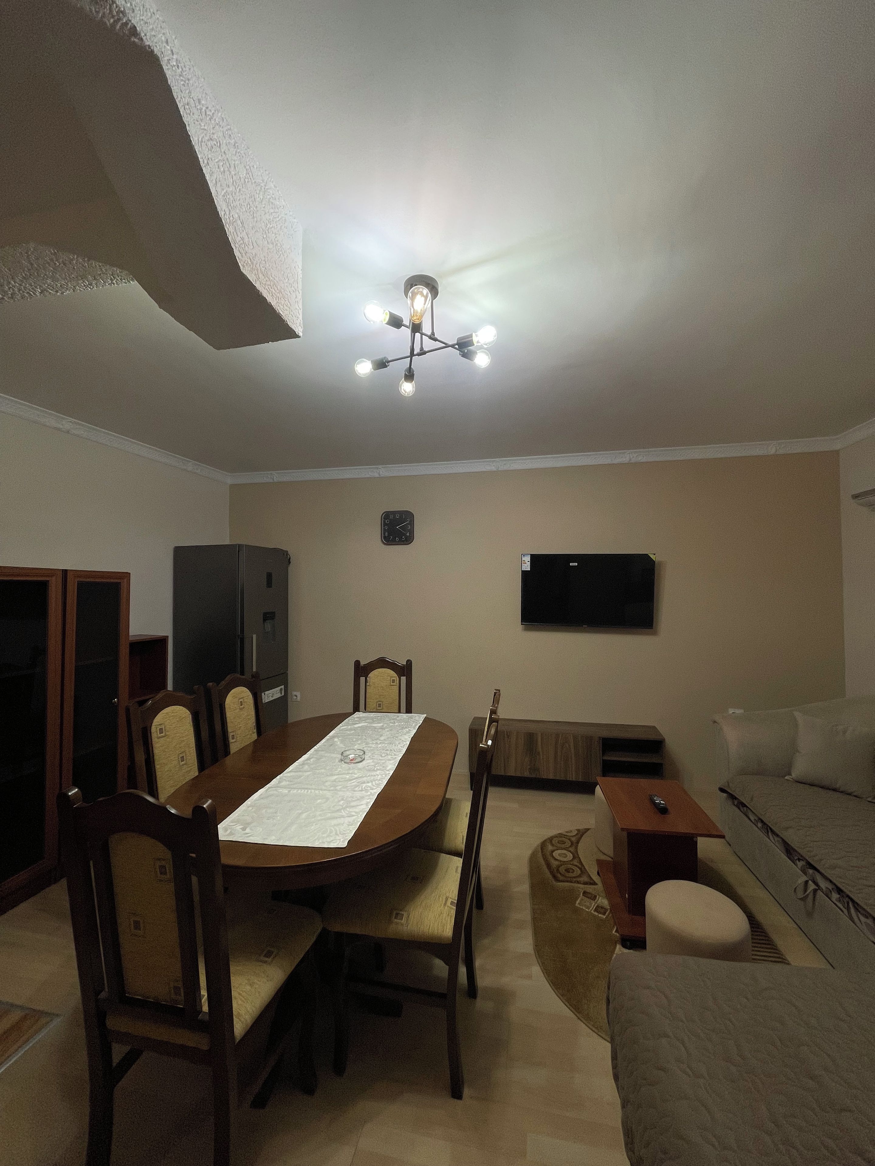 Uma sala de jantar com mesa de madeira, cadeiras, um lustre, uma televisão e um sofá.