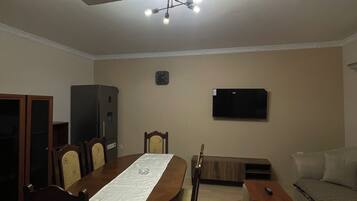 Uma sala de jantar com mesa de madeira, cadeiras, um lustre, uma televisão e um sofá.