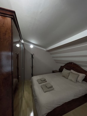 A bedroom with a sloped ceiling, a wooden wardrobe, a bed with pillows and folded towels, and a coat rack.