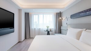 Ein Hotelzimmer mit einem großen Bett, einem flachbildfernseher an der Wand, einem kleinen Tisch mit einer Vase und einem fenster mit Vorhängen.