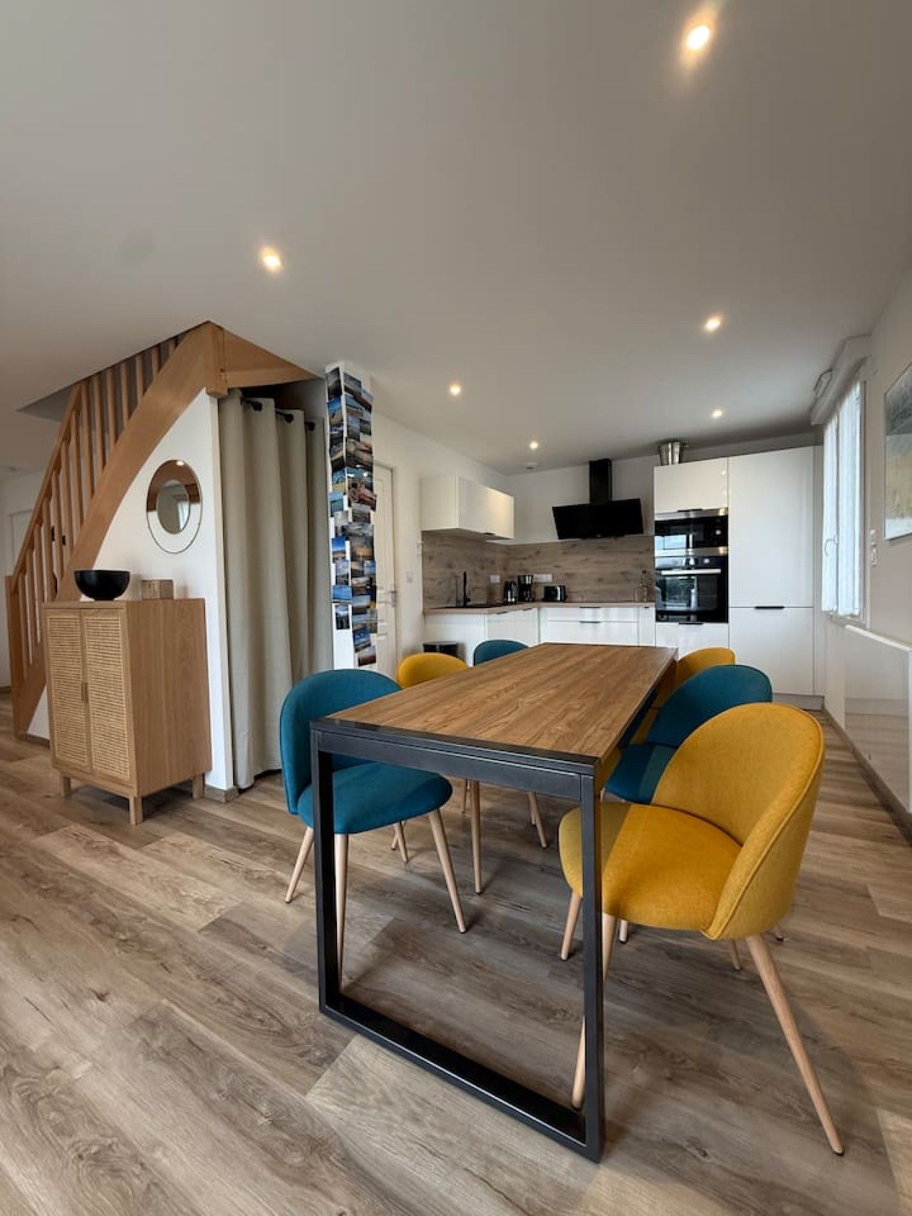 A modern kitchen with white cabinets, a wooden staircase, and a dining area with a wooden table and colorful chairs.