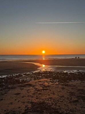 Ein Strand bei Sonnenuntergang, feuchter Sand und einige Personen in der Ferne.