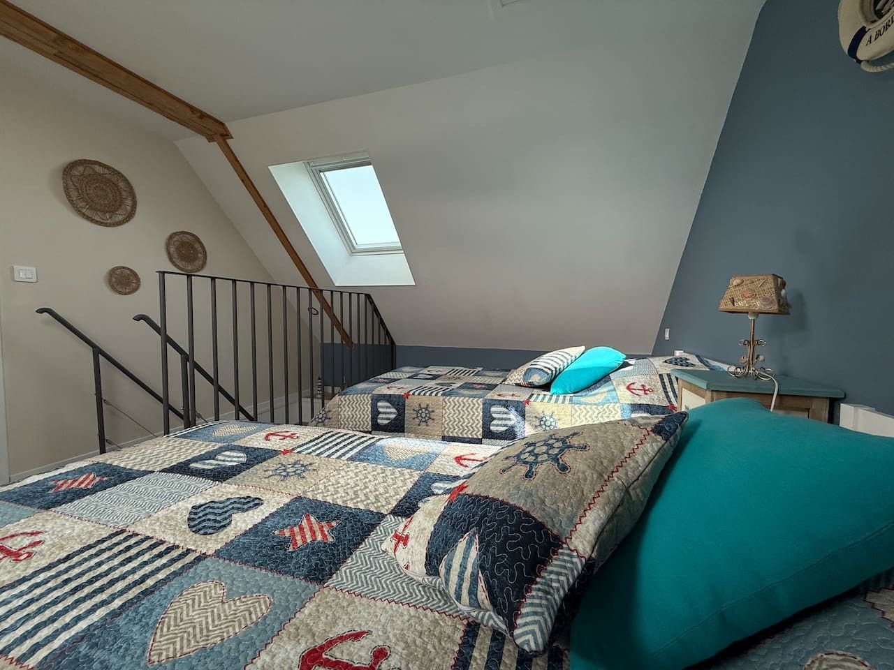 Ein Schlafzimmer mit schrägen Wänden, einem Oberlicht, einem Bett mit einem gemusterten Quilts, einem türkisfarbenen Kissen und einem kleinen Tisch mit einer Lampe.