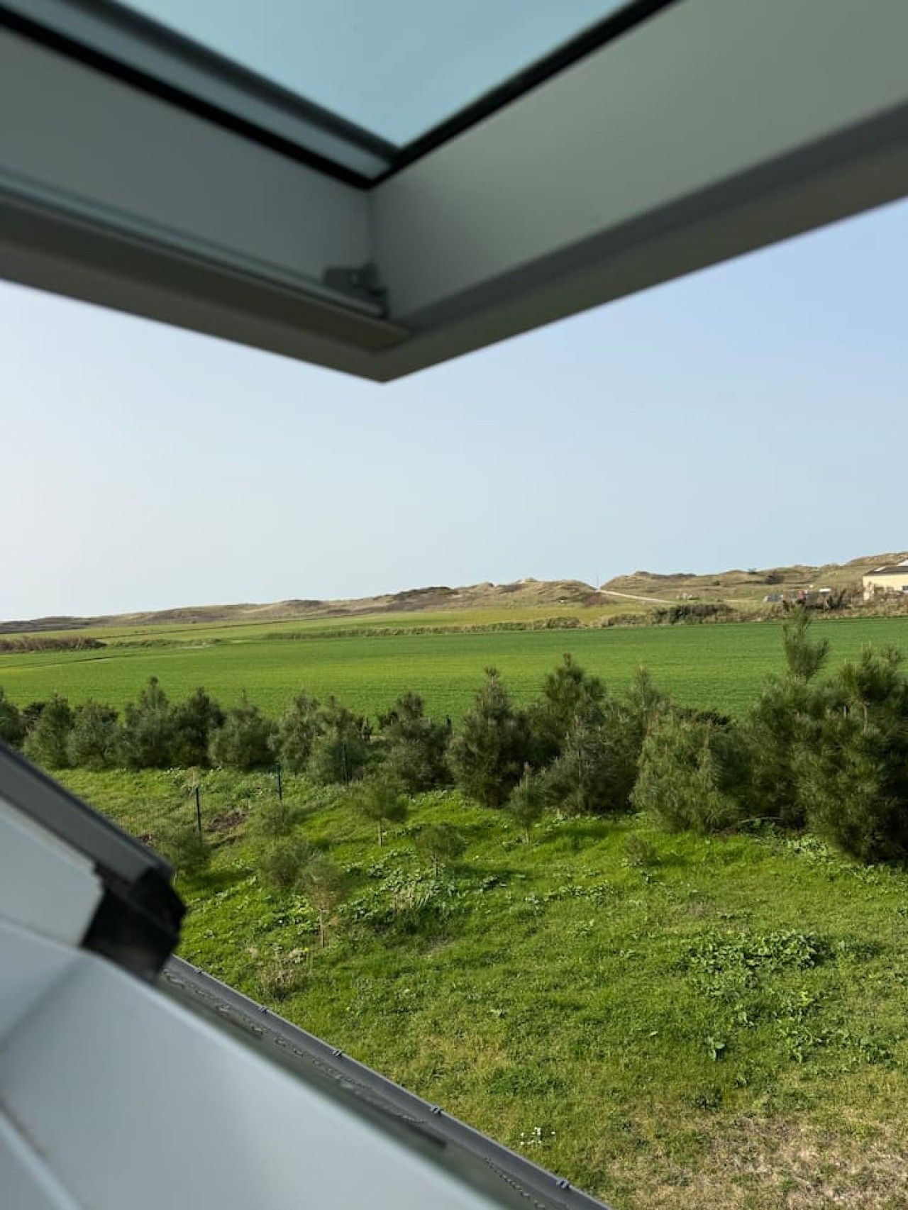 Blick aus einem Fenster auf einen klaren Himmel, ein grünes Feld und ferne Hügel.