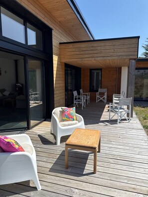 Eine moderne Außenterrasse mit Holzboden, weißen Möbeln und Glastüren, die zu einem Innenbereich führen.