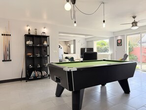 A modern indoor pool table with a green felt surface, surrounded by a black pool table base. There is a black shelving unit with numbered compartments against the wall, and a ceiling fan above. - Family home near the beach, 5 bedrooms, pool table, garden (Bretteville-sur-Ay)
