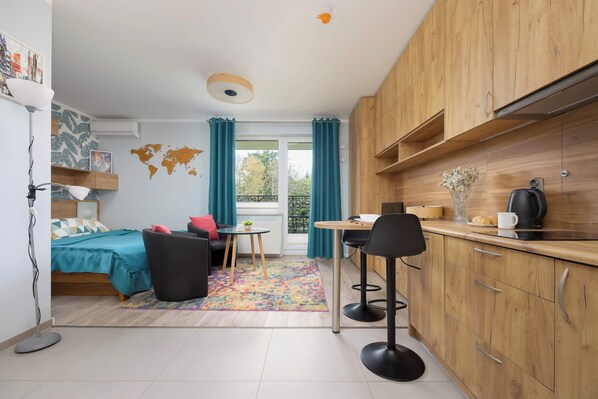 A compact room with a bed, a small sofa, a dining table with two chairs, a kitchenette, and a view through a window.