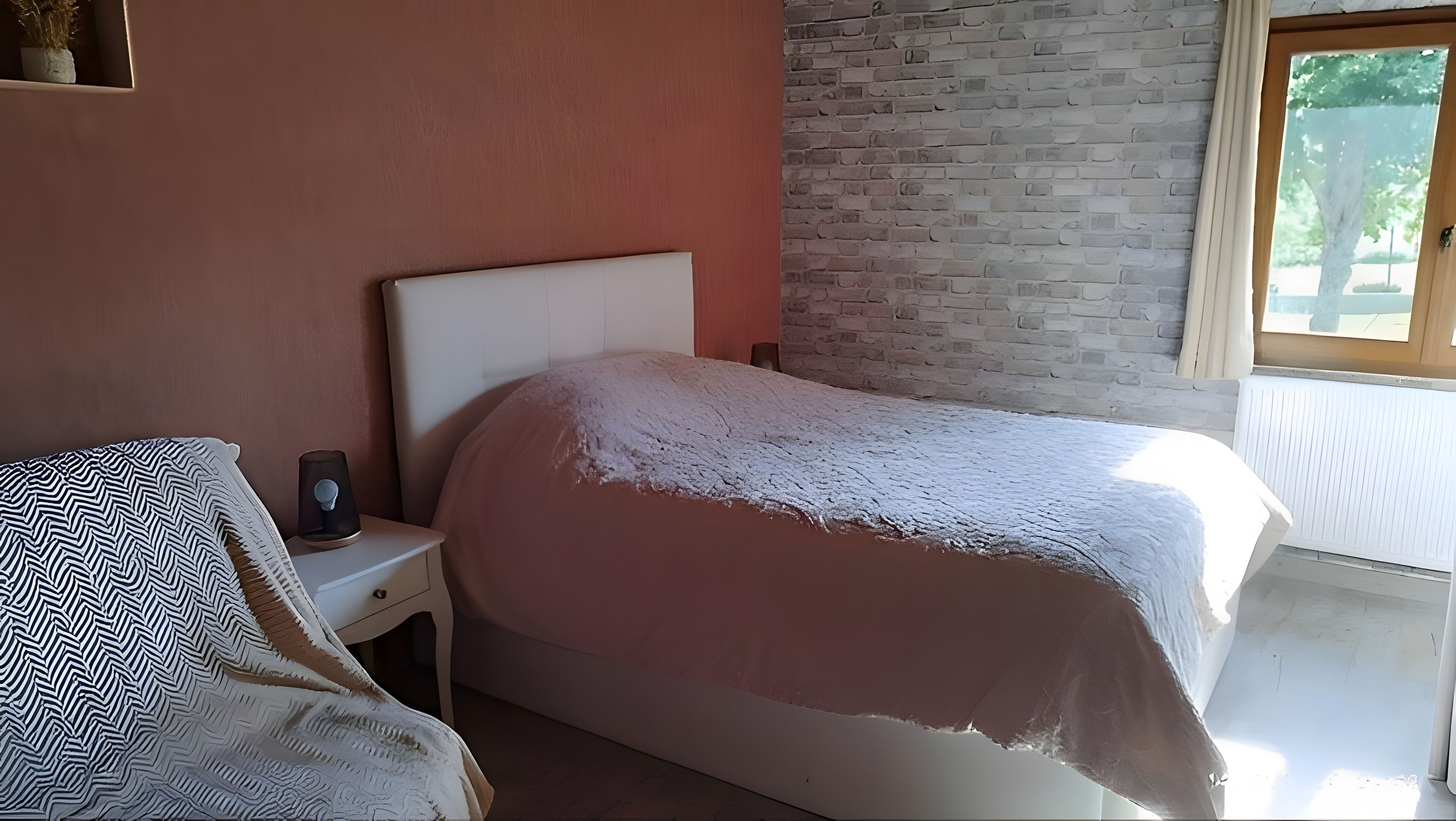 Quarto com cama, mesa de cabeceira, planta em vaso e janela.