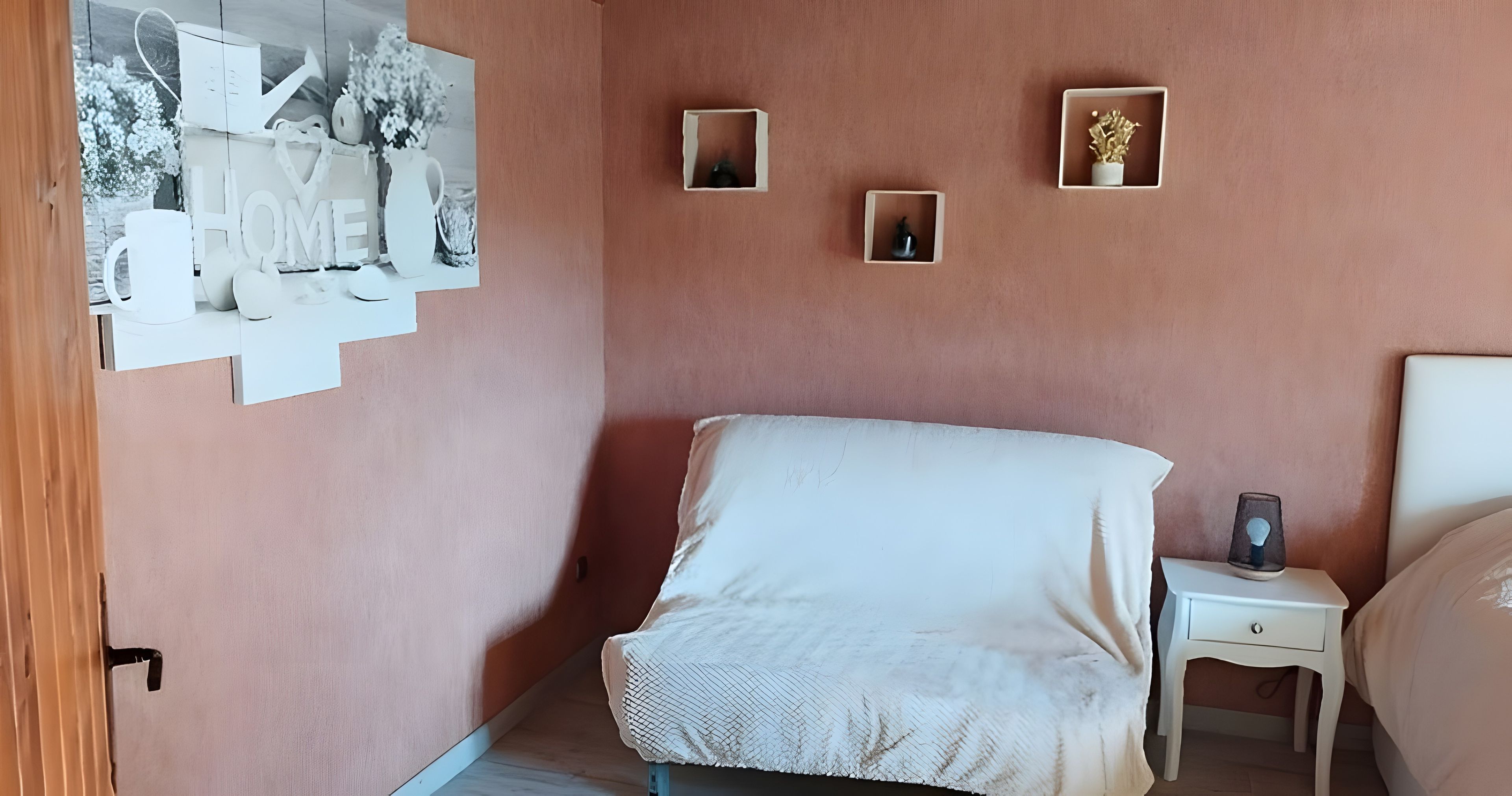 Un dormitorio con cama, una mesita de noche, un estante en la pared con objetos decorativos y una puerta con picaporte.