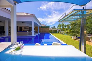 Ein moderner Poolbereich im Freien mit überdachter Sitzgelegenheit und Blick auf ein Haus mit einem Ziegeldach.