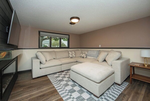 A living room with a sectional sofa, a small ottoman, a patterned rug, a window, and a ceiling light fixture.