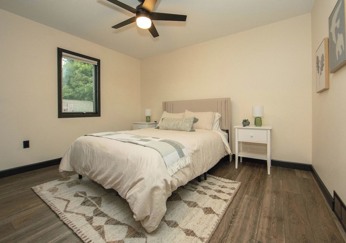 A bedroom with a bed, a nightstand, a bedside table, a ceiling fan, a window, and a wall-mounted picture.