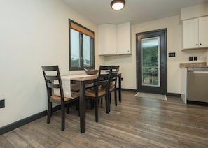 A dining area with a wooden table and chairs, a window with blinds, and a door leading to a kitchen with cabinets.