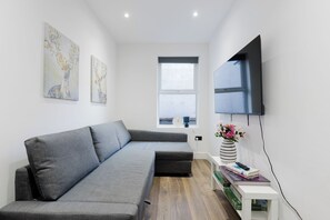 A modern living room with a grey sectional sofa, a flat-screen TV mounted on the wall, and a window with a view of the sky. - Modern 4BDR Flat in the Heart of Wandsworth (Greater London)
