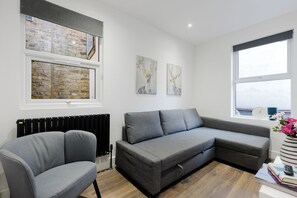 A modern living room with a grey sectional sofa, a grey armchair, and a radiator. There are two framed artworks on the wall, and a window with a view of the outside. - Modern 4BDR Flat in the Heart of Wandsworth (Greater London)
