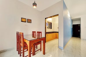 A dining area with a wooden table and chairs, a water dispenser, and a kitchen visible through an open doorway. - Super Hotel O Rose Garden Ooty (Ootacamund)