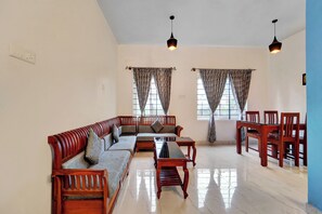A spacious living room with a wooden sofa set, a dining table with chairs, and two windows with curtains. - Super Hotel O Rose Garden Ooty (Ootacamund)