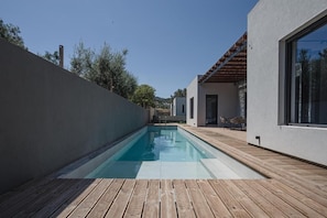 A modern outdoor swimming pool with a wooden deck, surrounded by a white wall and a building with large windows. - NIMA Suites (Palairos)
