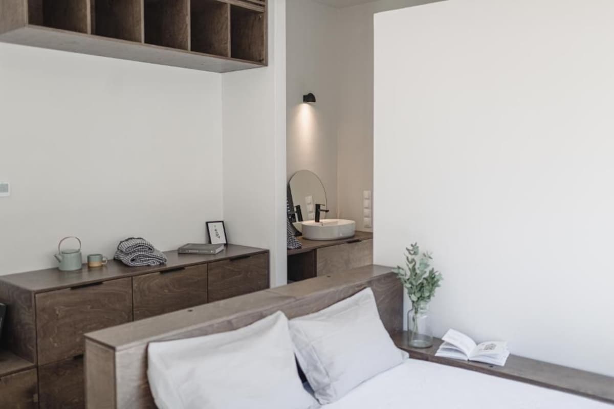 A modern bedroom with a wooden headboard, a bedside table with a book and a vase, and a large mirror above a white sink.