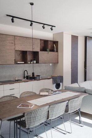 A modern kitchen with wooden cabinets, a dining area with a table and chairs, and a built-in oven. - NIMA Suites (Palairos)
