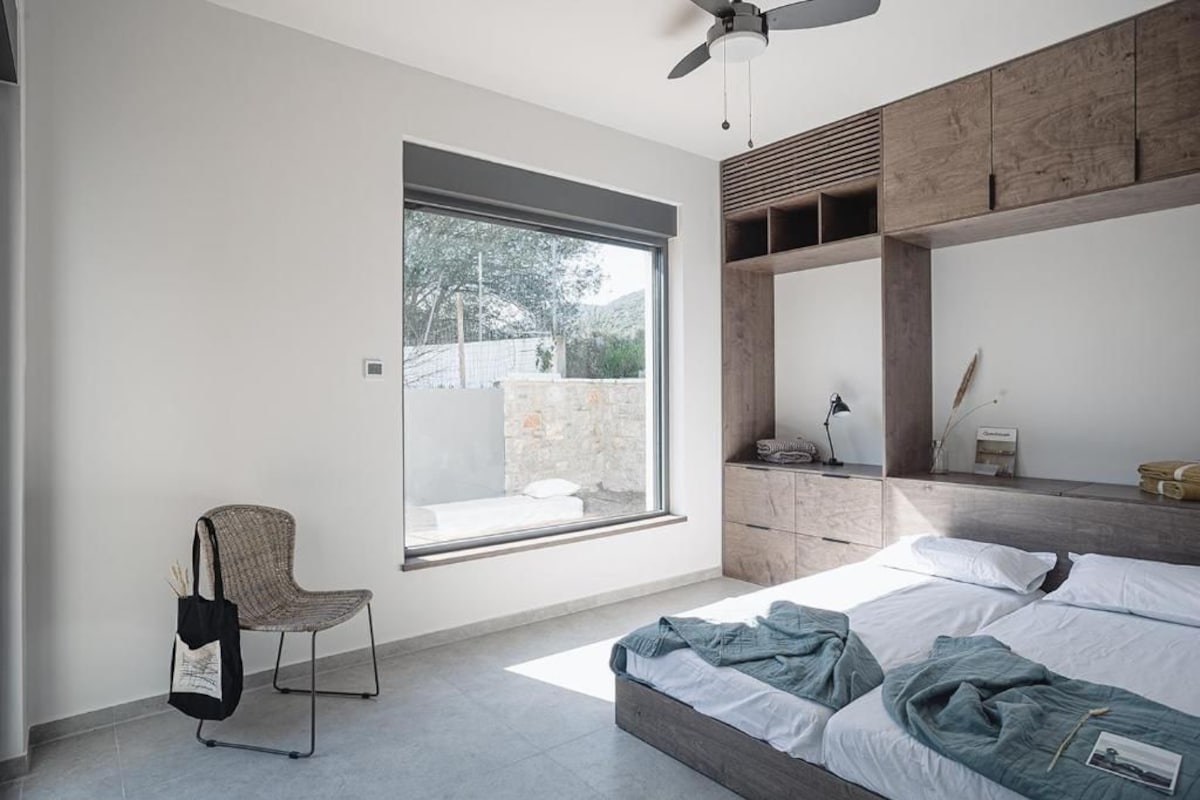 A bedroom with a large window, a bed, a chair, and a wooden cabinet.