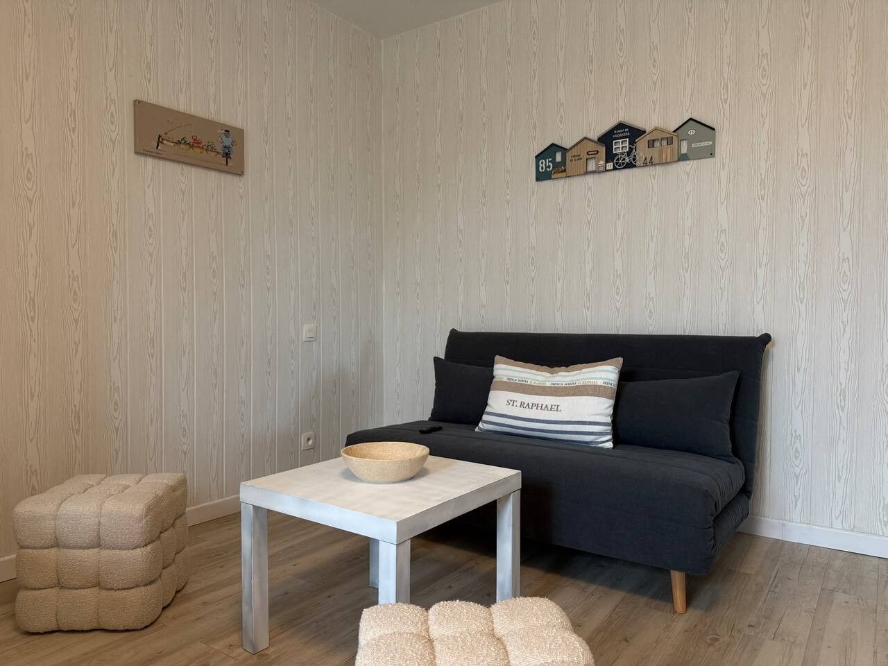 Ein Wohnzimmer mit einer dunklen Couch, einem weißen Tisch mit einer Schüssel, einem gepolsterten Hocker und einem Wandregal mit hausförmigen Dekorationen.