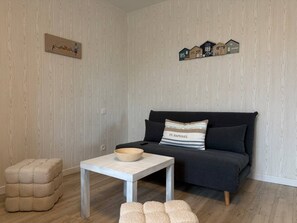A living room with a dark sofa, a white table with a bowl, a cushioned stool, and a wall-mounted shelf with house-shaped decorations. - La Plava, a 1-minute walk from the ocean, Créances (Créances)