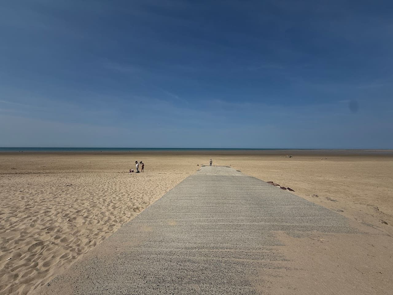 Una playa con un camino de hormigón que lleva al agua, cielo despejado y algunas personas a lo lejos.