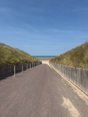 A paved pathway leading to a beach, flanked by wooden fences and grassy dunes. - La Plava, a 1-minute walk from the ocean, Créances (Créances)