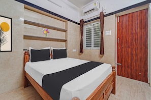 Um quarto com cama, guarda-roupa de madeira, ar condicionado, janela com cortinas e prateleira na parede com flores.