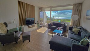 Una sala de estar moderna con un televisor de pantalla plana, dos sofás, una mesa de centro y un amplio ventanal corredero que da a un balcón con vistas al mar.