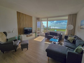A modern living room with a flat-screen TV, two sofas, a coffee table, and a large sliding glass door leading to a balcony with a view of the sea. - Vrbo Property (Großenbrode)