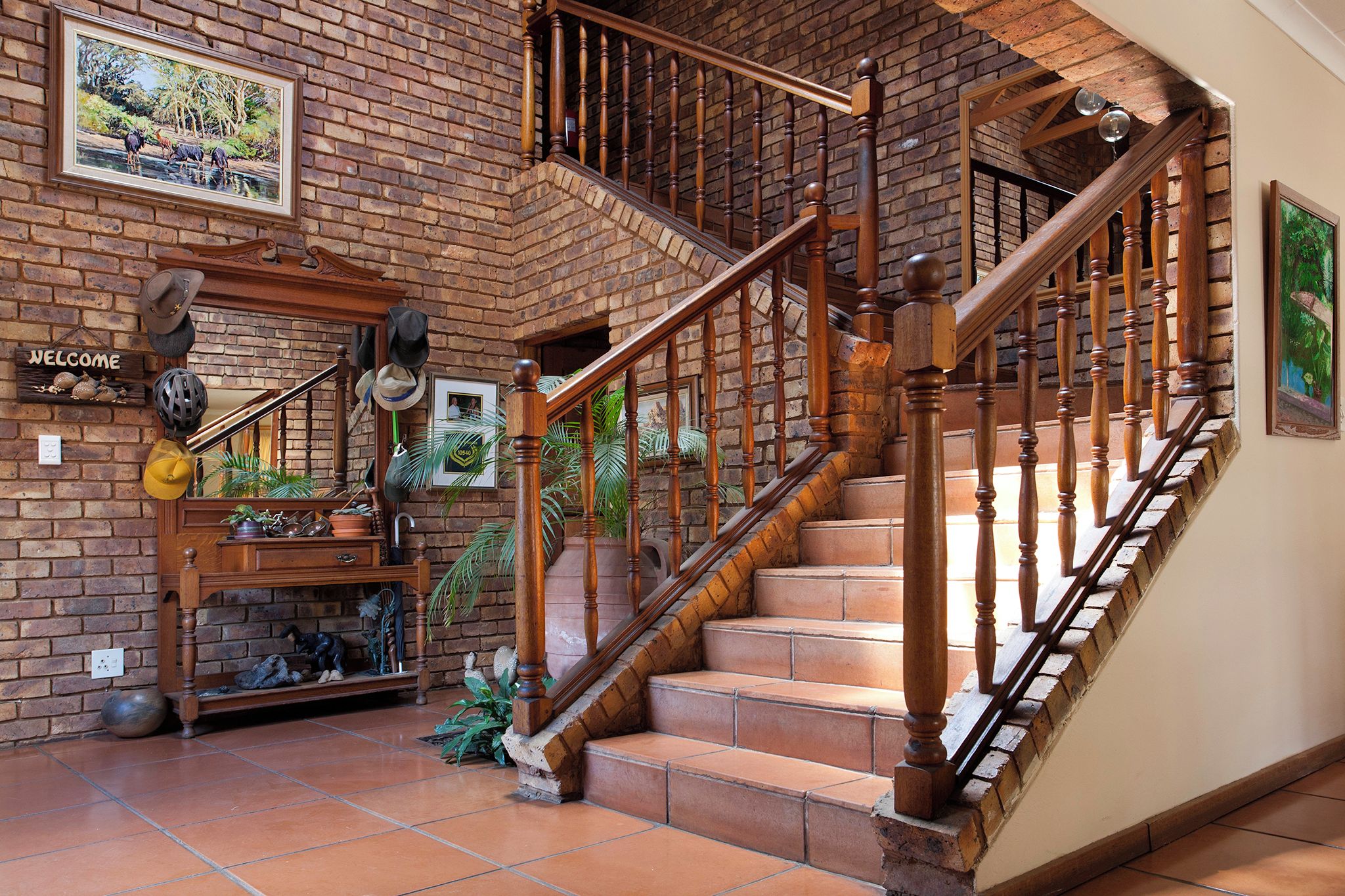 Una escalera de madera con un fondo de pared de ladrillo, un letrero de bienvenida y varios objetos decorativos, incluyendo sombreros y un cuadro enmarcado.
