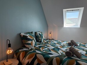 Ein Schlafzimmer mit schrägverdachter Decke, einem Bett mit blattgemusterten Bettwäsche, einer Nachttischlampe, einem Fenster mit Blick und zusammengerollten Handtüchern auf dem Bett.