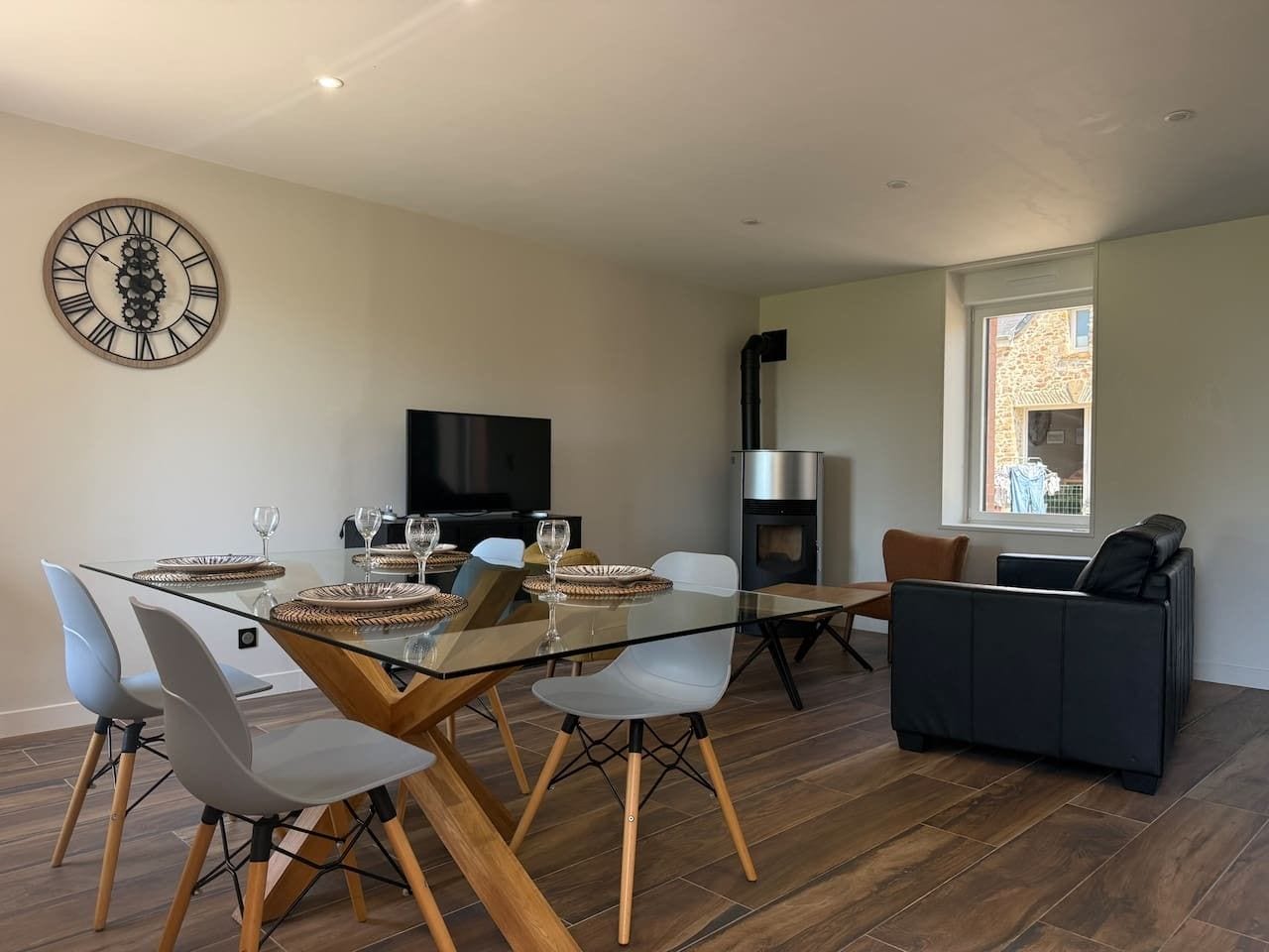 A modern living space with a glass dining table, comfortable seating, a fireplace, and a large wall clock.