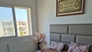 A bedroom with a bed, a nightstand, a framed certificate on the wall, and a window with a view of buildings. - Vrbo Property (Monastir)