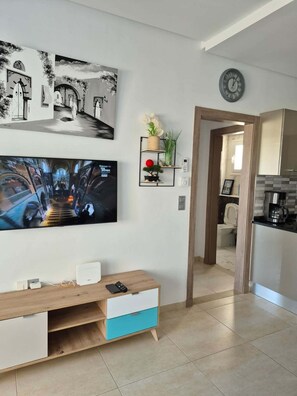 A modern living room with a flat-screen TV, a wooden entertainment unit, and a kitchen area visible through an open doorway.