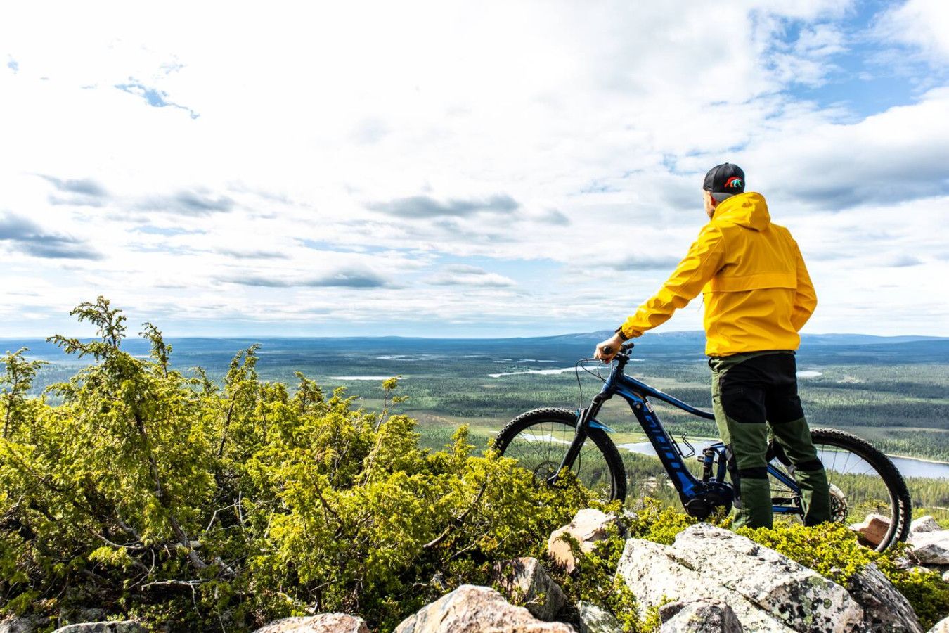 Una persona de pie junto a una bicicleta de montaña sobre terreno rocoso, con vista a un paisaje boscoso.