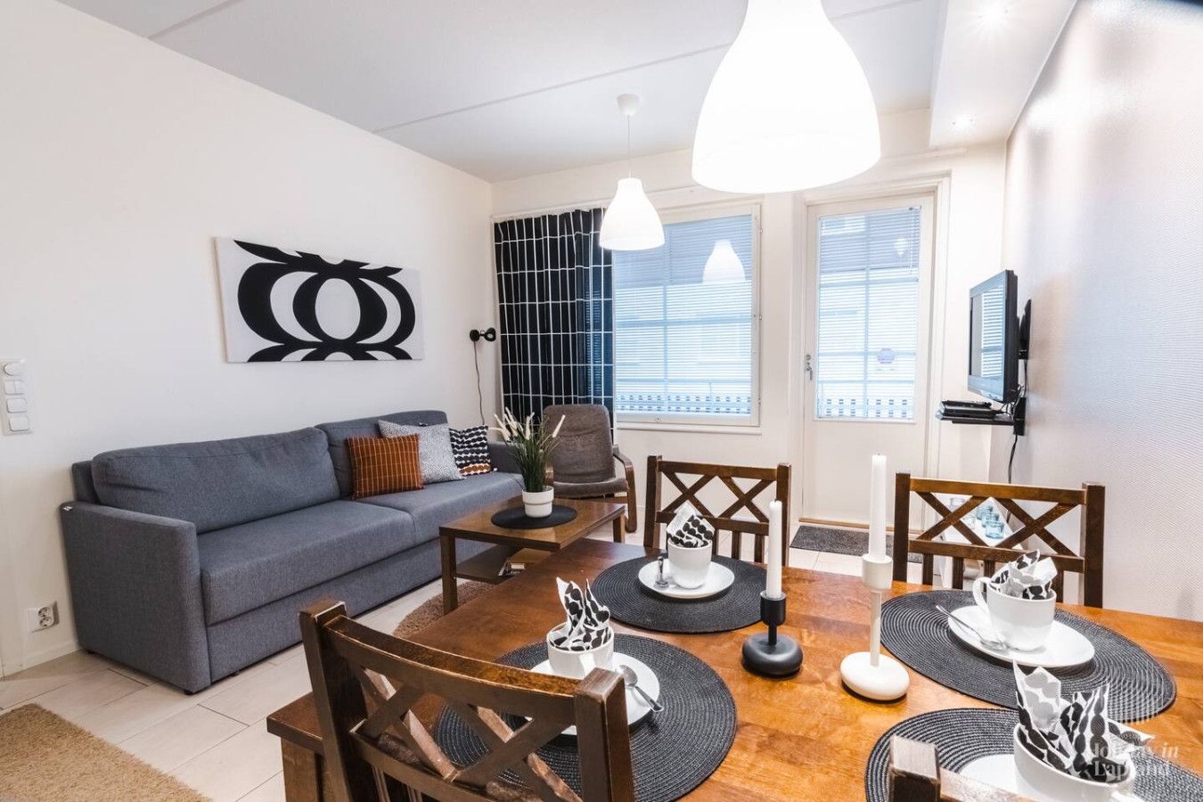 A modern living room with a grey sofa, wooden dining table, and chairs. There is a TV mounted on the wall and a window with blinds.