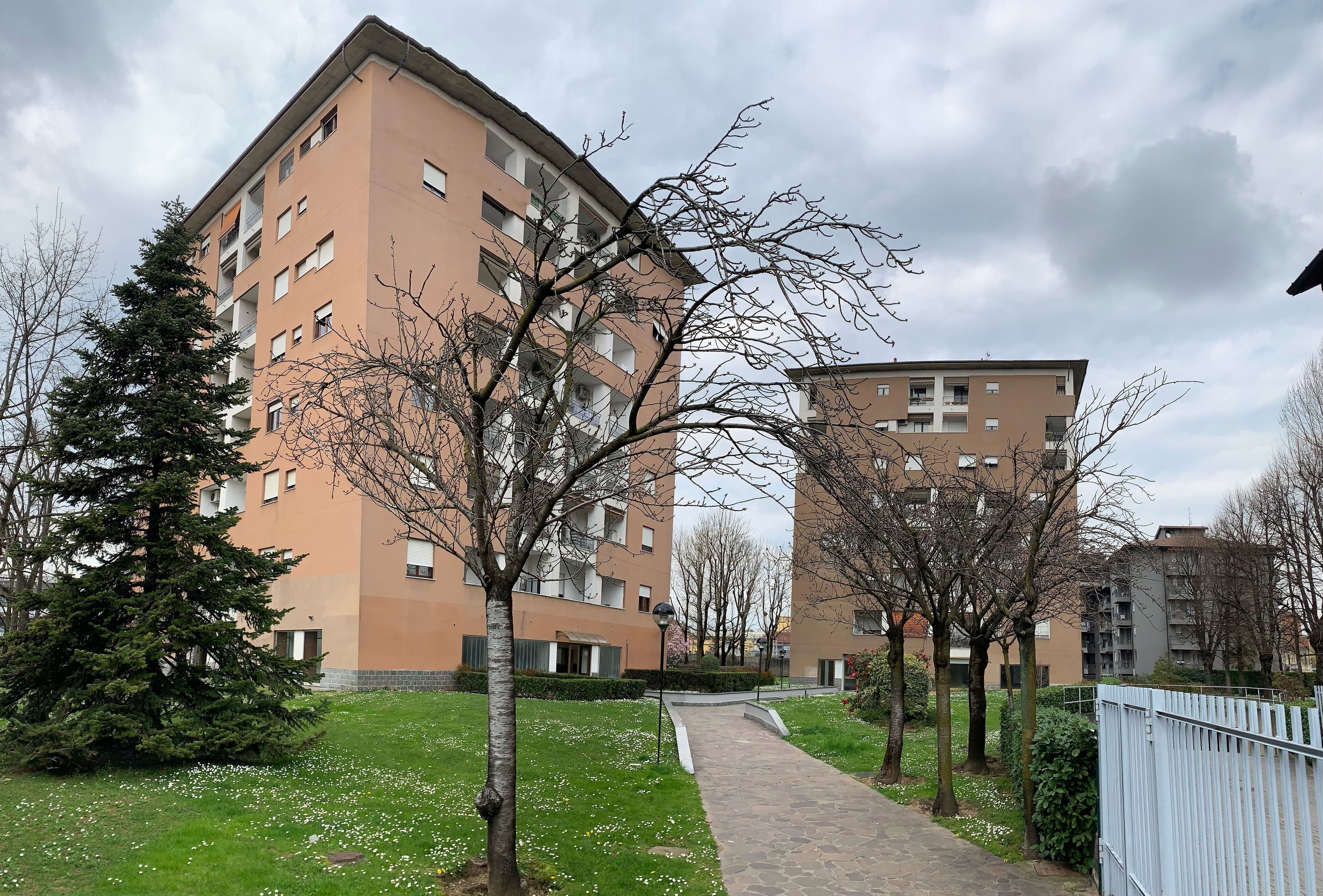 Un edificio residencial de varios pisos, un sendero pavimentado y árboles con follaje escaso.
