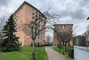 Un edifici residencial amb diversos pisos, una passera pavimentada i arbres amb fulles escasses.