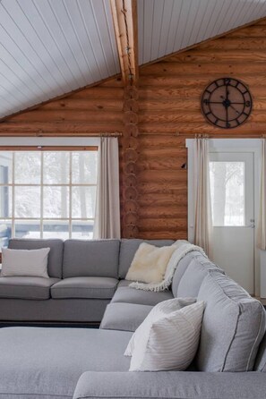 Ein Holzblockhaus-Interieur mit einer Eckcouch, einer Holzbalkendecke und einem großen Fenster.