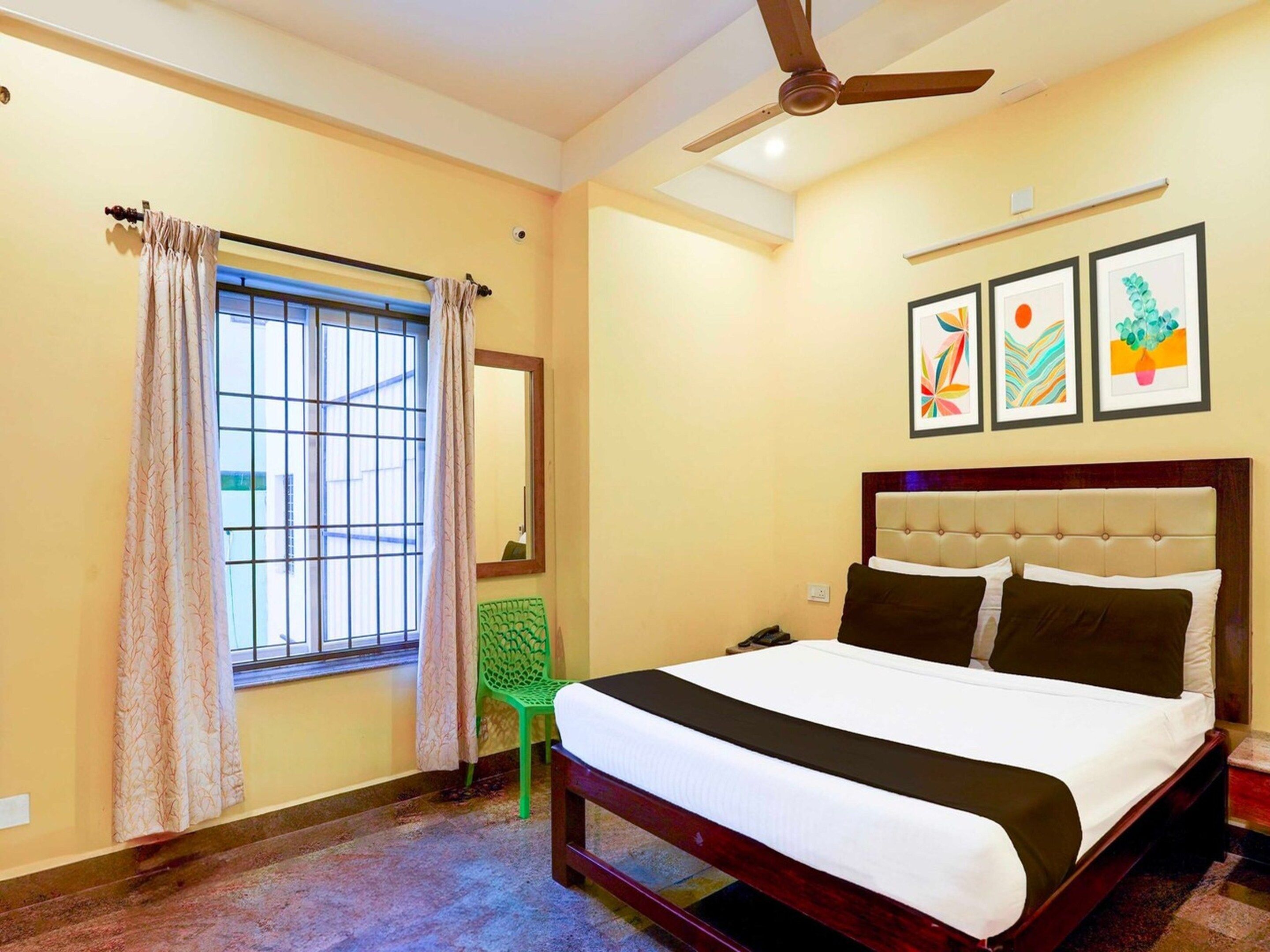 A hotel room with a bed, a ceiling fan, a window with curtains, a green chair, and three framed artworks on the wall.