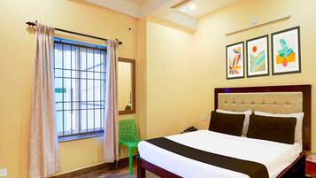 A hotel room with a bed, a ceiling fan, a window with curtains, a green chair, and three framed artworks on the wall.