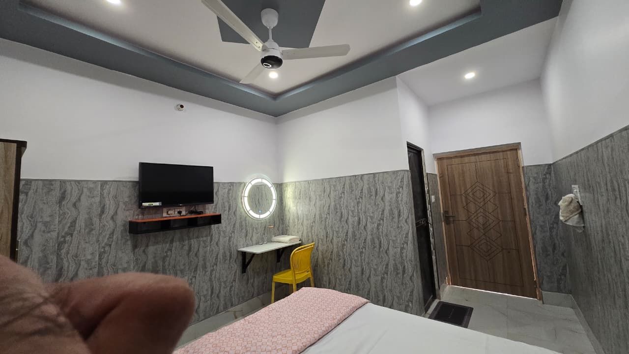 A hotel room with a bed, a desk with a chair, a television, a ceiling fan, and a wooden door.