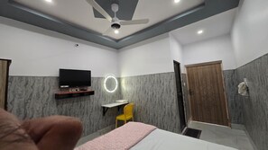 A hotel room with a bed, a desk with a chair, a television, a ceiling fan, and a wooden door.